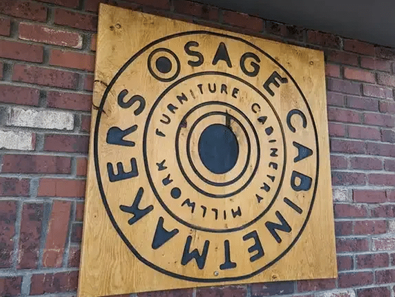 A wooden sign mounted on a brick wall. The sign features a circular design with bold black lettering that reads "Osage Cabinet Makers, Furniture, Cabinetry, Millwork" arranged in concentric circles.