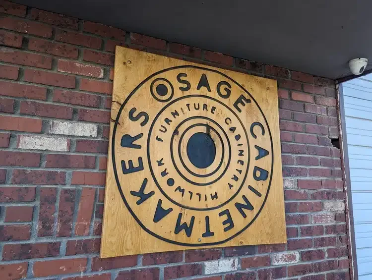 A wooden sign mounted on a brick wall featuring the text "Osage Cabinet Makers" in bold black letters. The sign includes a circular design with an inner circle and the words "Furniture" and "Cabinet" integrated within it, against a yellow background.
