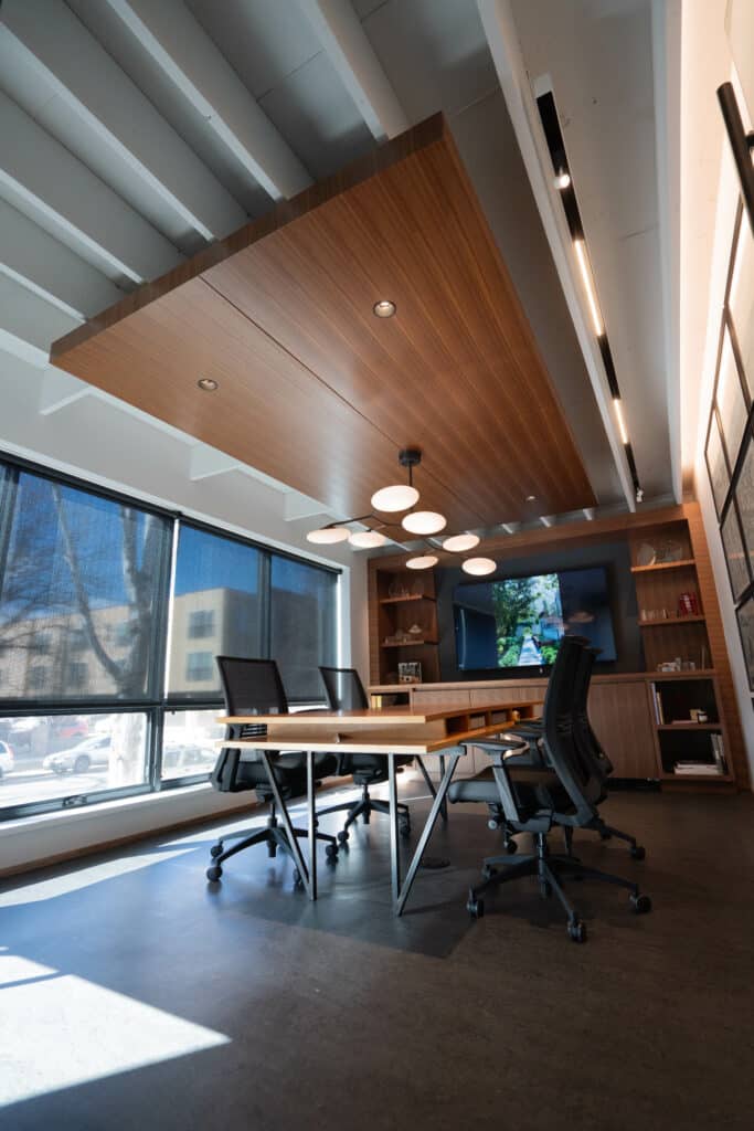 A modern conference room featuring a large wooden table surrounded by black chairs. Above the table, a wooden ceiling panel with integrated lighting hangs. Large windows allow natural light to fill the space, and a TV screen is mounted on the wall.