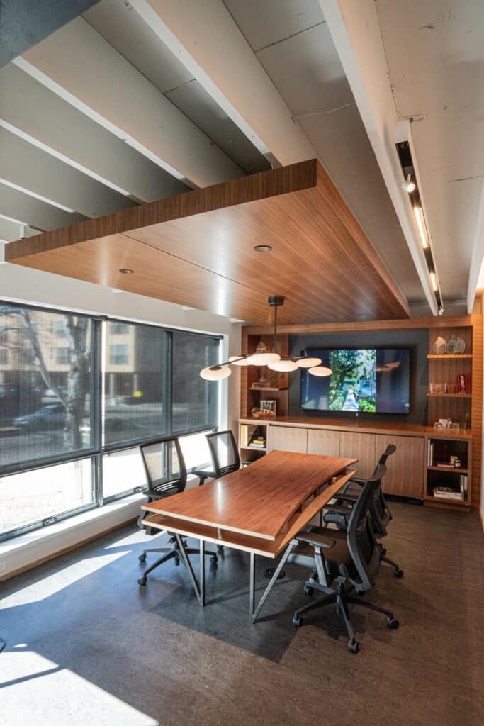 A modern conference room featuring a long wooden table with ergonomic black chairs. Large windows allow natural light to enter, enhancing the space. A wall-mounted screen displays an image, and wooden paneling gives a warm finish.