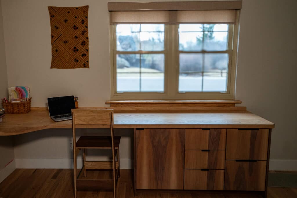 A minimalistic home office setup featuring a wooden desk with a light-colored surface and three drawers, a wooden chair, and a laptop in a basket on the left. A large window provides natural light, with a textured wall hanging above the desk.