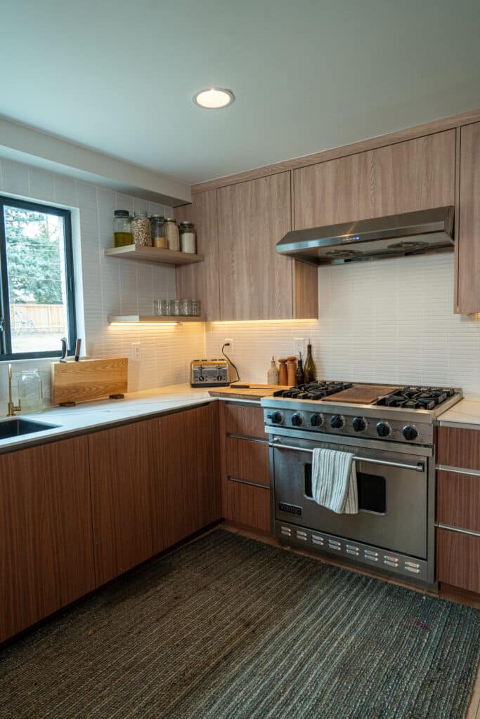 A modern kitchen featuring wooden cabinetry, a stainless steel gas stove, and a hood. There is a sink under a window, a wooden cutting board, and a toaster on the countertop. Soft lighting highlights the space, which has a textured rug on the floor.