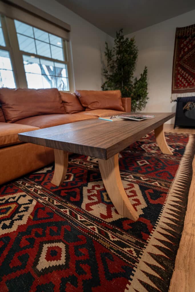 A wooden coffee table with tapered legs sits in front of a brown leather sofa, on a patterned area rug. Natural light comes through large windows, and a green plant is visible in the background.