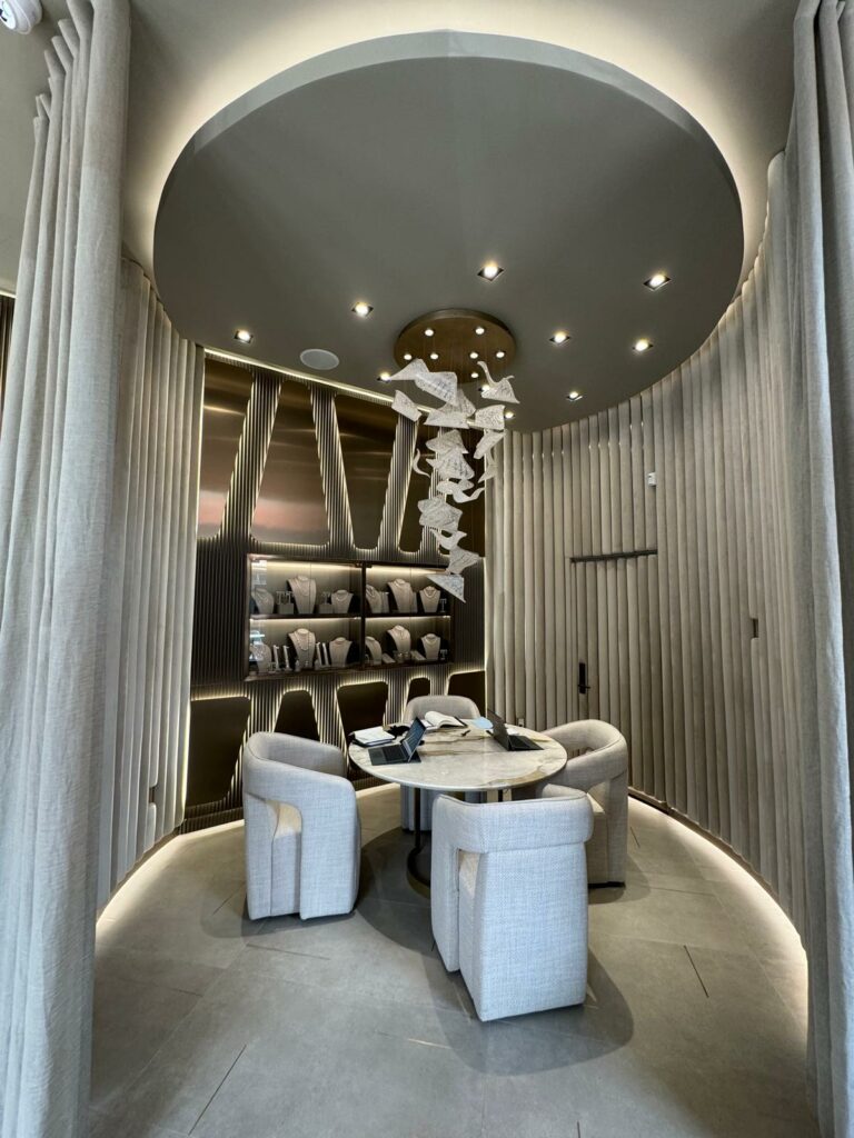 A modern, circular meeting room with a domed ceiling. The walls are lined with vertical panels. In the center, a round table is surrounded by four light-colored chairs. Shelves display various items, and a decorative mobile hangs from the ceiling.