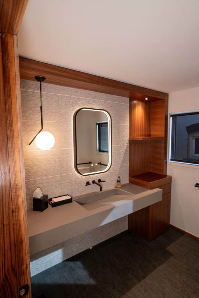 A modern bathroom featuring a rectangular sink with a minimal design. A round mirror is mounted on a textured wall above the sink. A pendant light hangs from the ceiling, and the cabinet has warm wood tones, while a window allows natural light.