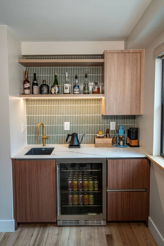 A modern kitchen corner featuring a wooden cabinet and an open shelf. The shelf displays various bottles and jars. Below is a small beverage fridge stocked with drinks, a black sink, and a kettle on the countertop. The space is well-lit with neutral tones.