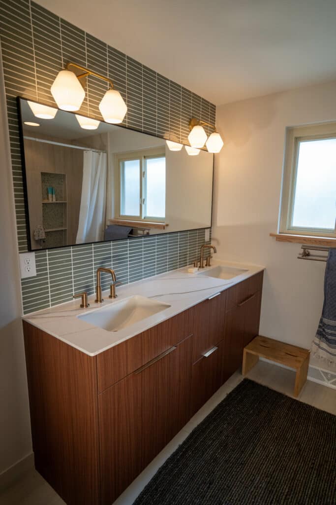 A modern bathroom featuring a double sink vanity with wooden cabinetry and a white countertop. Above the sinks, there is a large mirror and several light fixtures. The wall behind the sink is covered in green tiles, and a window provides natural light.