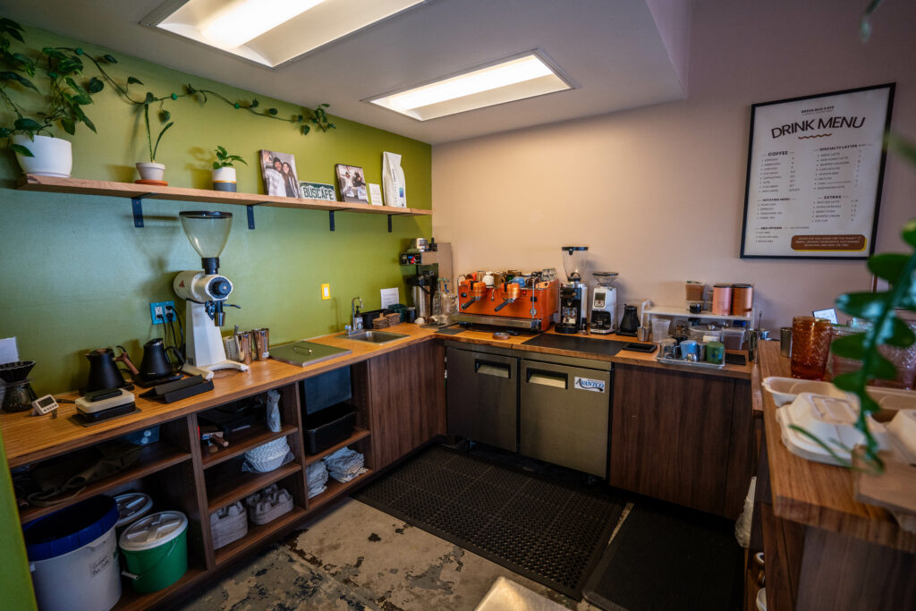 Coffee shop interior featuring a counter with espresso machines and coffee equipment. Shelves are stocked with cups and plants. A drink menu is displayed on the wall, showcasing available beverages. The floor shows a mix of concrete and tiles.