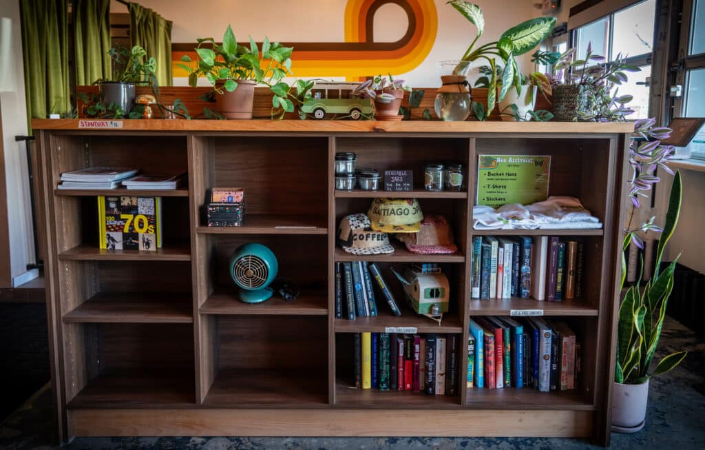 A wooden bookshelf displaying various items including books, magazines, potted plants, and decorative items like a vintage fan. The section on the left holds magazines, while the right has colorful books arranged neatly. Several plants are placed on top.