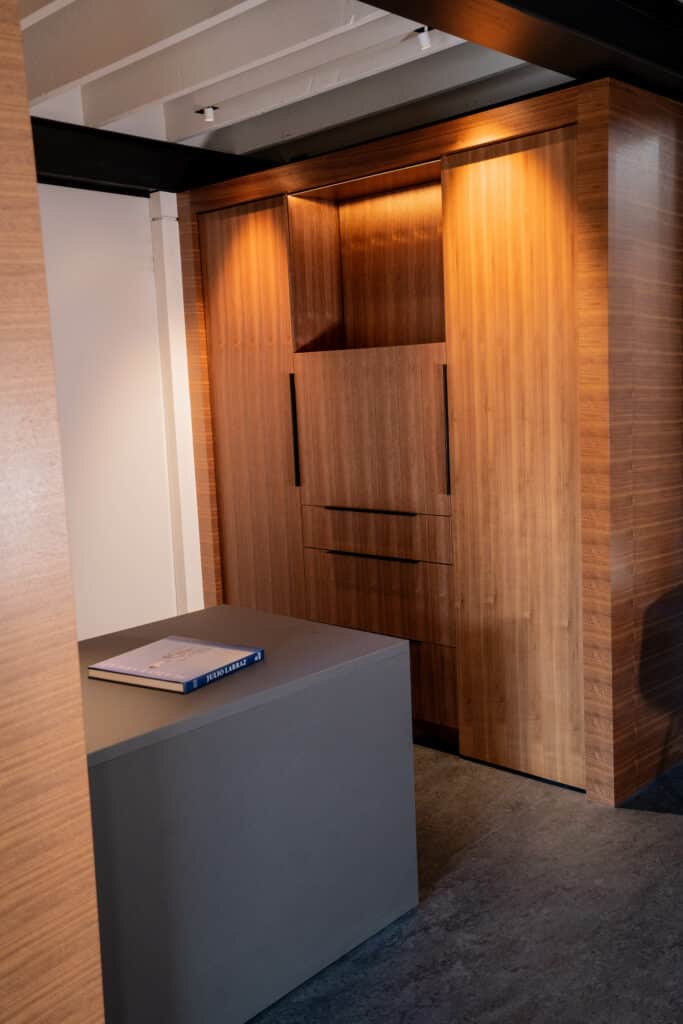 A modern interior space featuring a wooden storage unit with illuminated elements and a minimalist gray table in the foreground. The walls are light-colored, creating a contrast with the wooden surfaces.