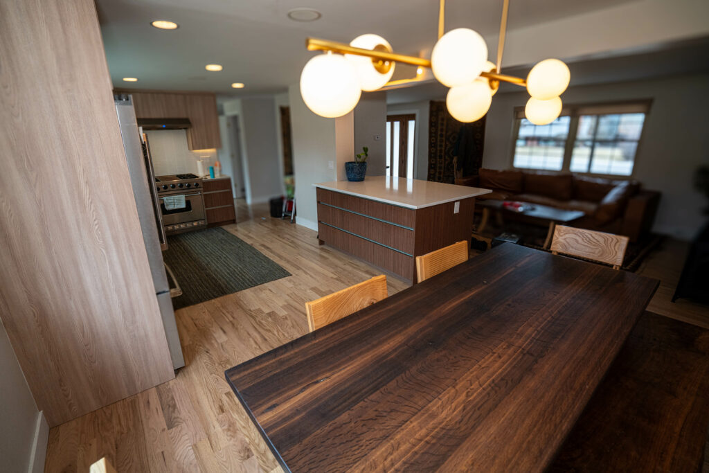A modern kitchen and dining area featuring a wooden dining table with four chairs, pendant lighting above, an island with a white countertop, and cabinets. Soft lighting illuminates the space, which has wooden floors and a view of the living room.