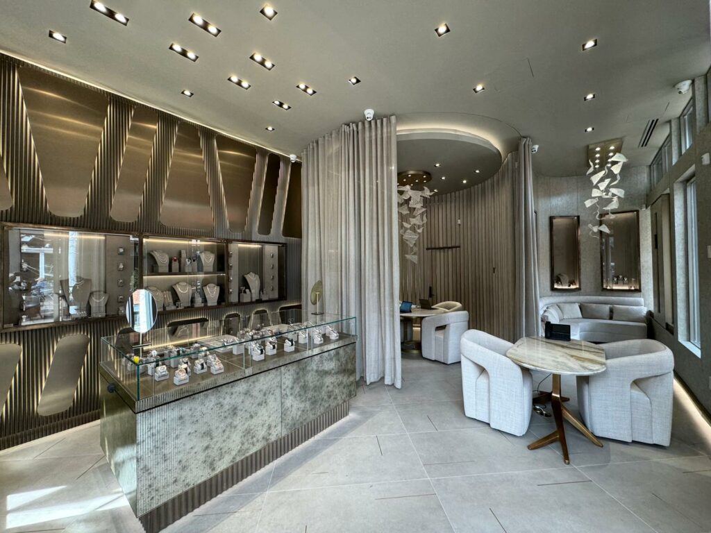 A modern jewelry store interior features a glass display case with various pieces of jewelry. The space is well-lit, with sleek wall designs and beige seating areas. Curtains flow between sections, and stylish decor elements are visible on the walls.