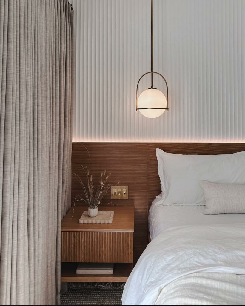 A cozy bedroom featuring a wooden bedside table with a small vase of dried plants, a white bed with pillows, and a hanging pendant light. Soft, textured curtains frame the window, complementing the neutral color scheme and subtle lighting.