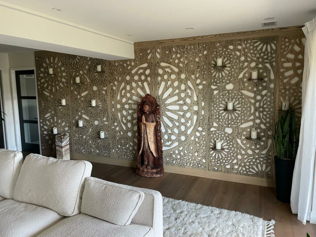 A living room featuring a decorative wall with intricate designs and several candle holders. In front of the wall is a wooden statue and a potted plant. A light-colored, plush sofa is positioned in the foreground.
