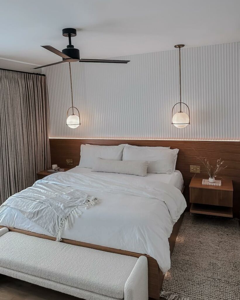 A contemporary bedroom featuring a large bed with white bedding, two round pendant lights, and a wooden headboard. A beige bench is placed at the foot of the bed, and light curtains, along with a ceiling fan, enhance the modern decor.